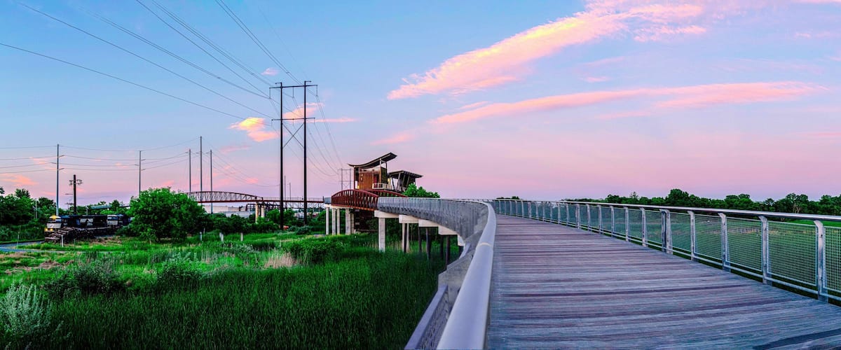 A wooden boardwalk with metal railings, a train track, and a bridge in the background.