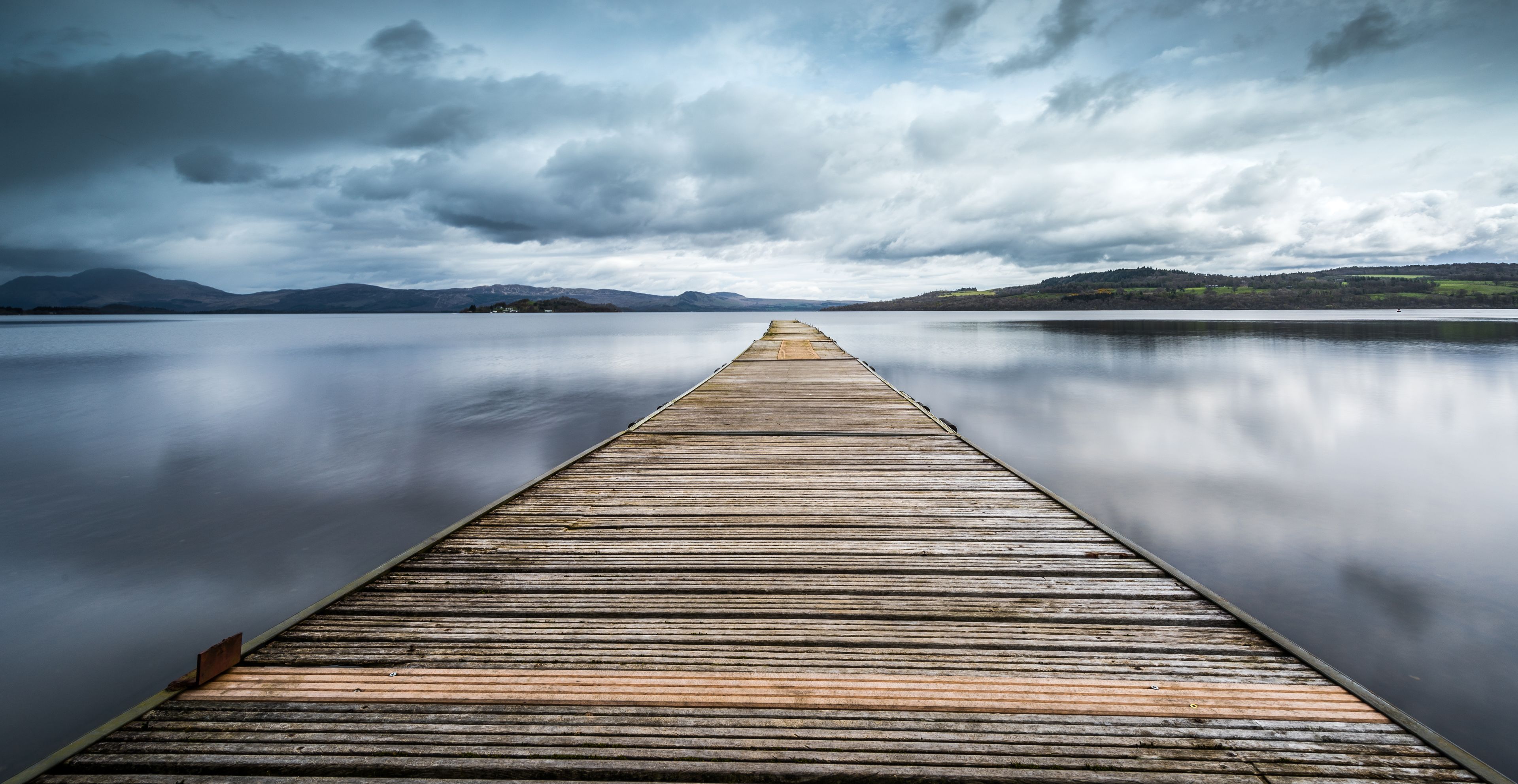 Un muelle de madera se extiende sobre aguas tranquilas, con montañas al fondo bajo un cielo nublado.