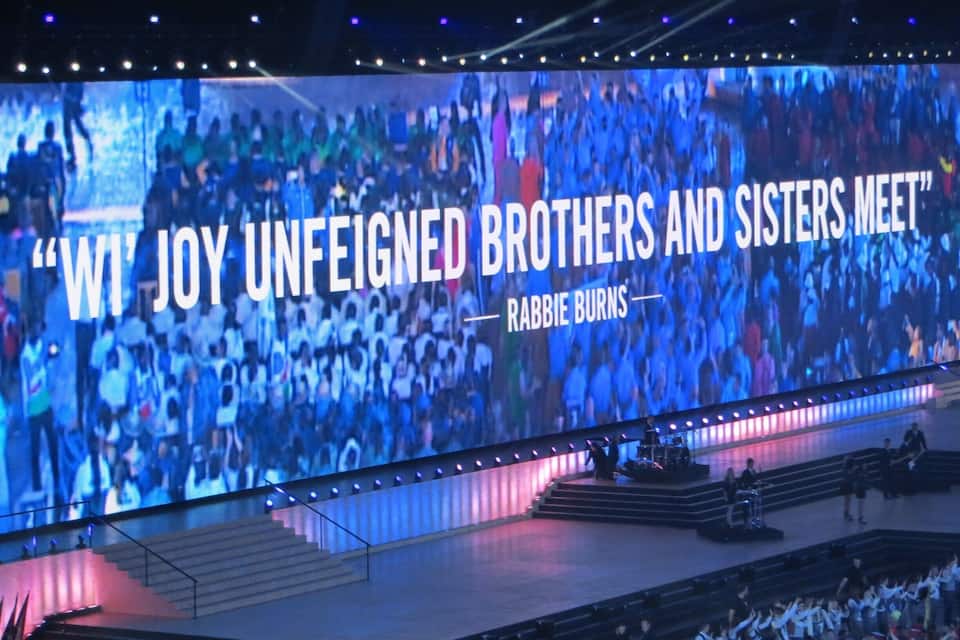 Eine Bühne mit einer großen Leinwand, auf der der Text „WI JOY UNFEIGNED BROTHERS AND SISTERS MEET“ von Rabie Burns steht.