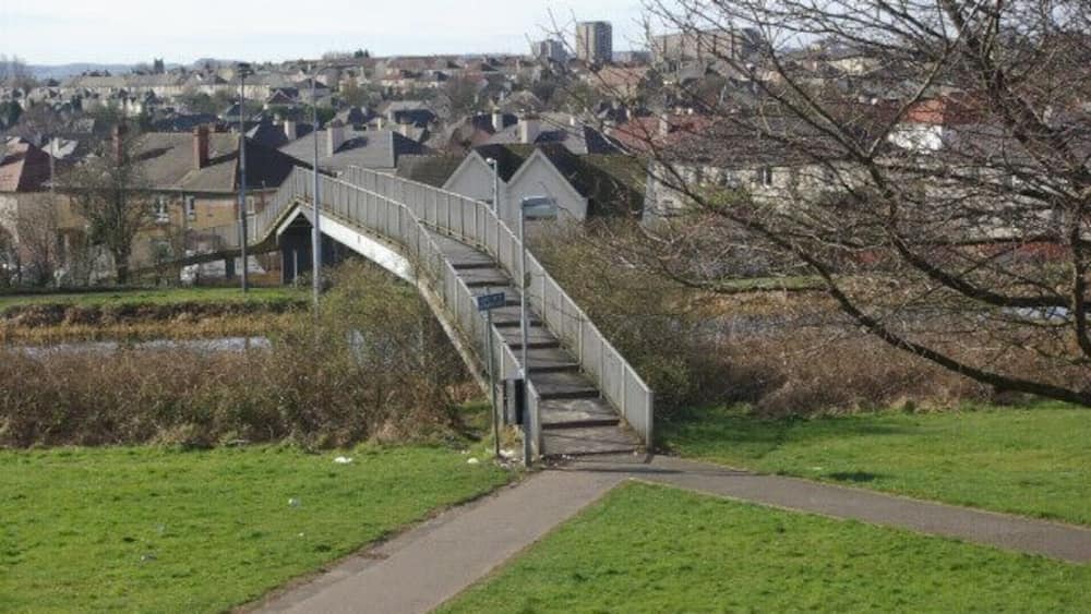 Un pont piétonnier enjambant une rivière, entouré d’herbe verte et bordé d’un sentier.