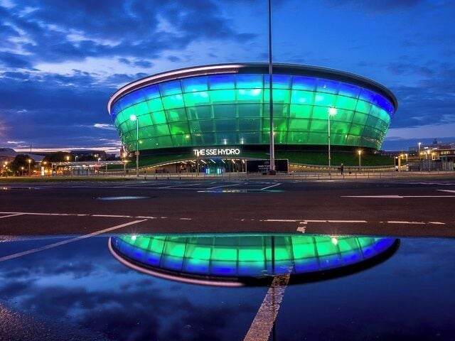 A modern stadium with a green and blue illuminated facade, surrounded by a wet reflective surface.