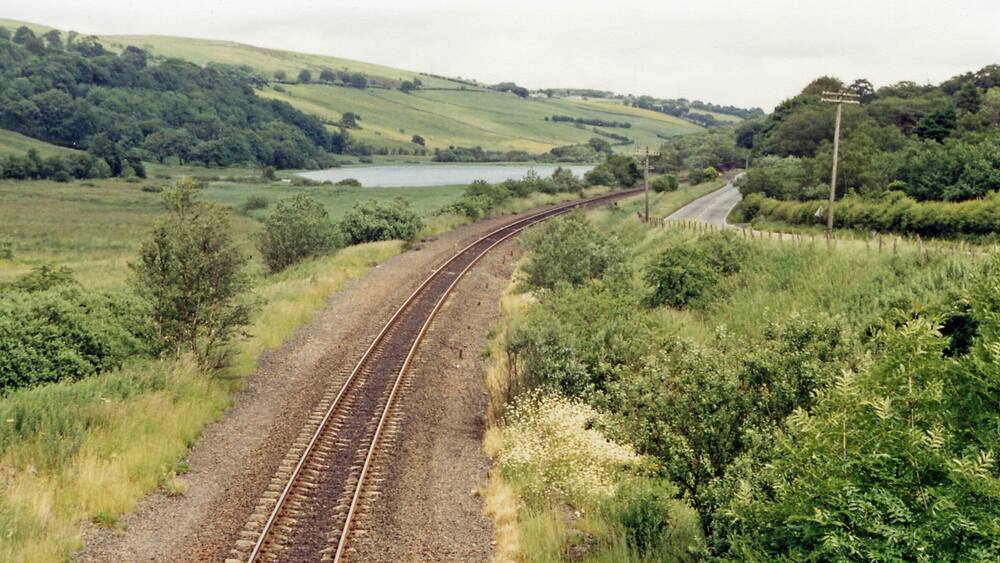 A railway track curving through a green landscape with hills and a lake in the distance.