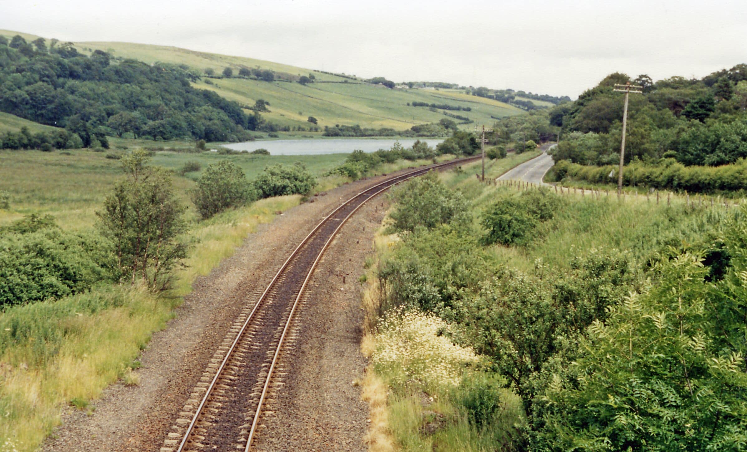 A railway track curving through a green landscape with hills and a lake in the distance.