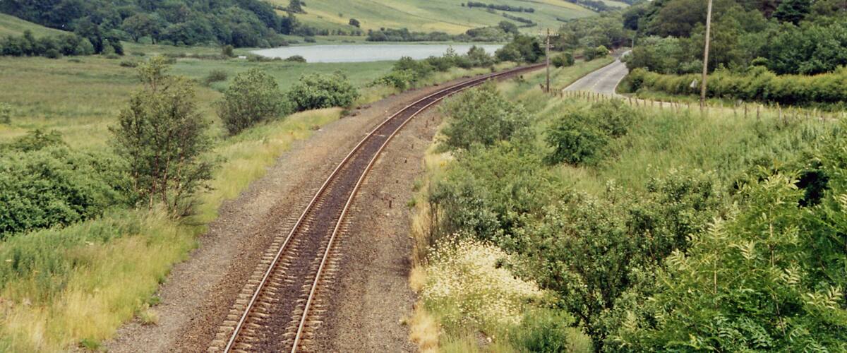 A railway track curving through a green landscape with hills and a lake in the distance.