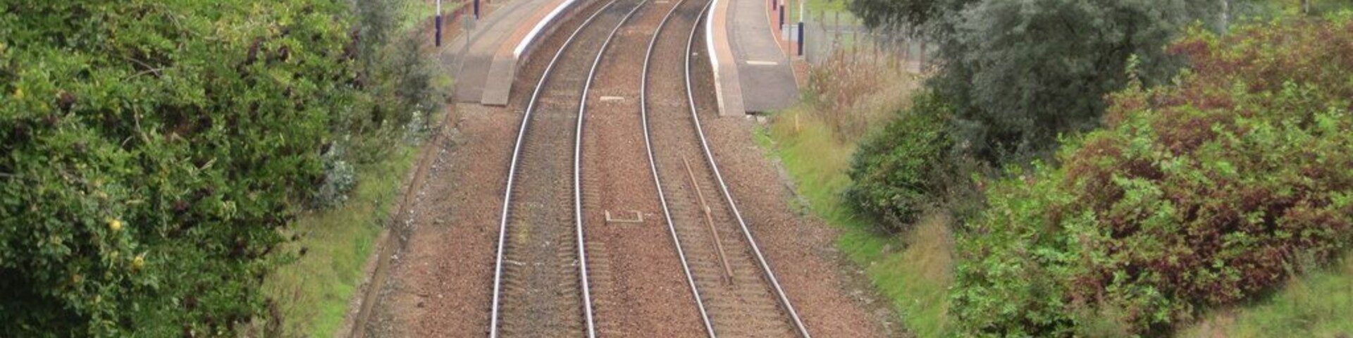 Railway tracks curving through a green landscape with trees and a building in the background.