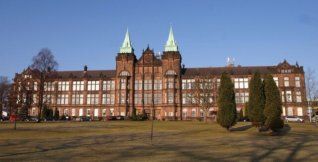 A large, historic building with two prominent green spires, surrounded by a grassy field and a few trees.