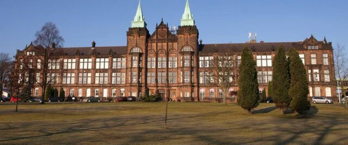 A large, historic building with two prominent green spires, surrounded by a grassy field and a few trees.