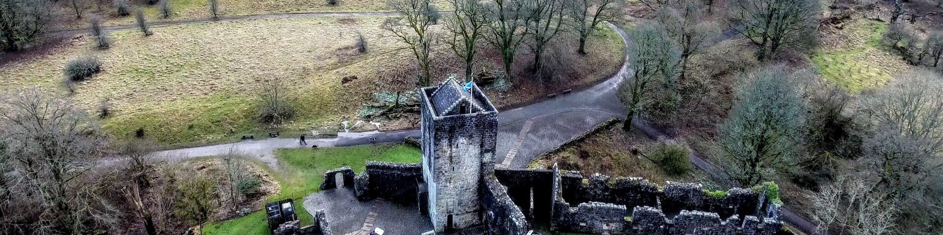 Vista aérea de um sítio histórico com ruínas, uma torre de vigia e uma estrada sinuosa por uma paisagem rural.