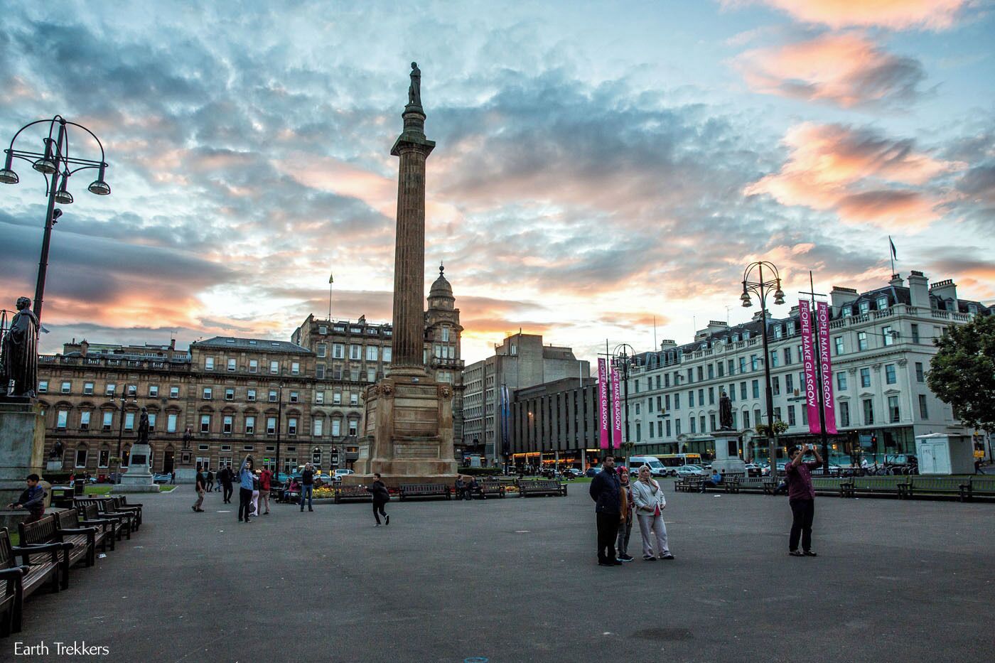 Uma praça urbana com um monumento imponente, edifícios históricos e um céu ao pôr do sol.