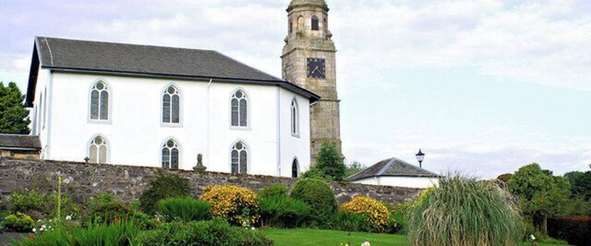 Une église blanche dotée d’un clocher, entourée d’un jardin bien entretenu où poussent diverses plantes et fleurs.