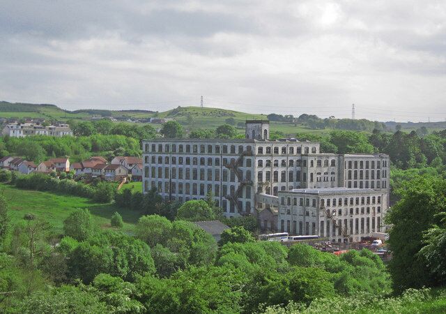 A large, multi-story building with a classical architectural style, surrounded by lush green fields and scattered houses.
