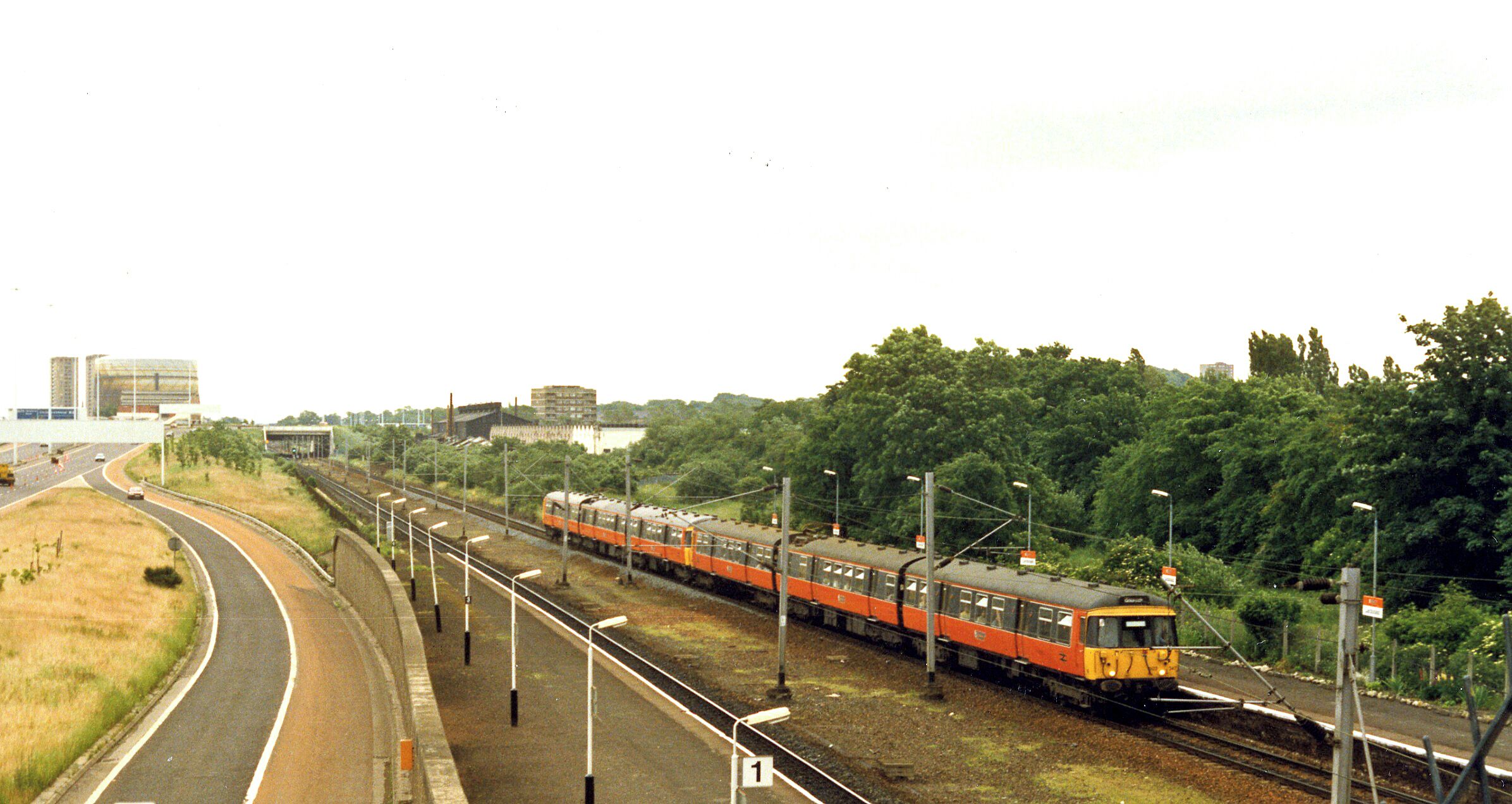 A train on tracks with a road curving to the left, surrounded by greenery and buildings in the background.