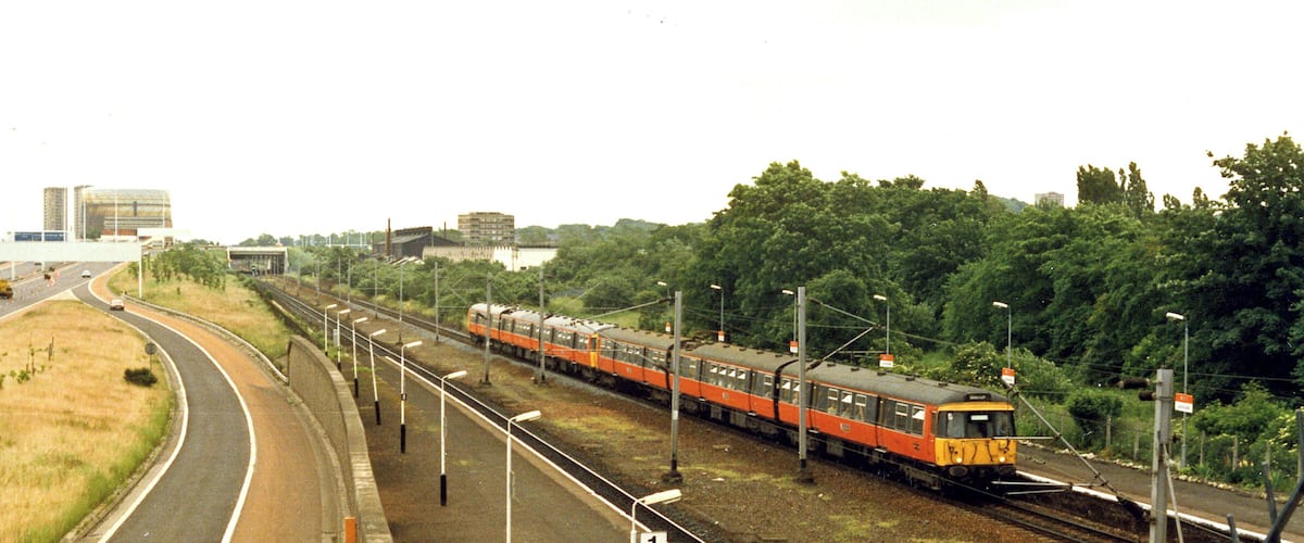 A train on tracks with a road curving to the left, surrounded by greenery and buildings in the background.