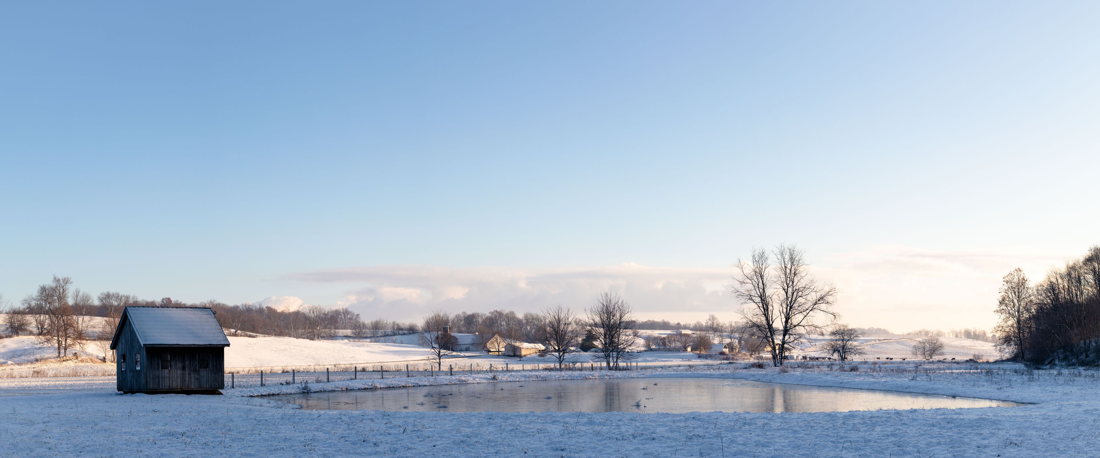 A snow-covered landscape with a small wooden structure, a pond, and bare trees.