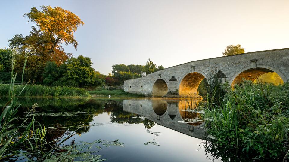 A stone bridge with three arches over a calm river, surrounded by lush greenery and trees with autumn foliage.