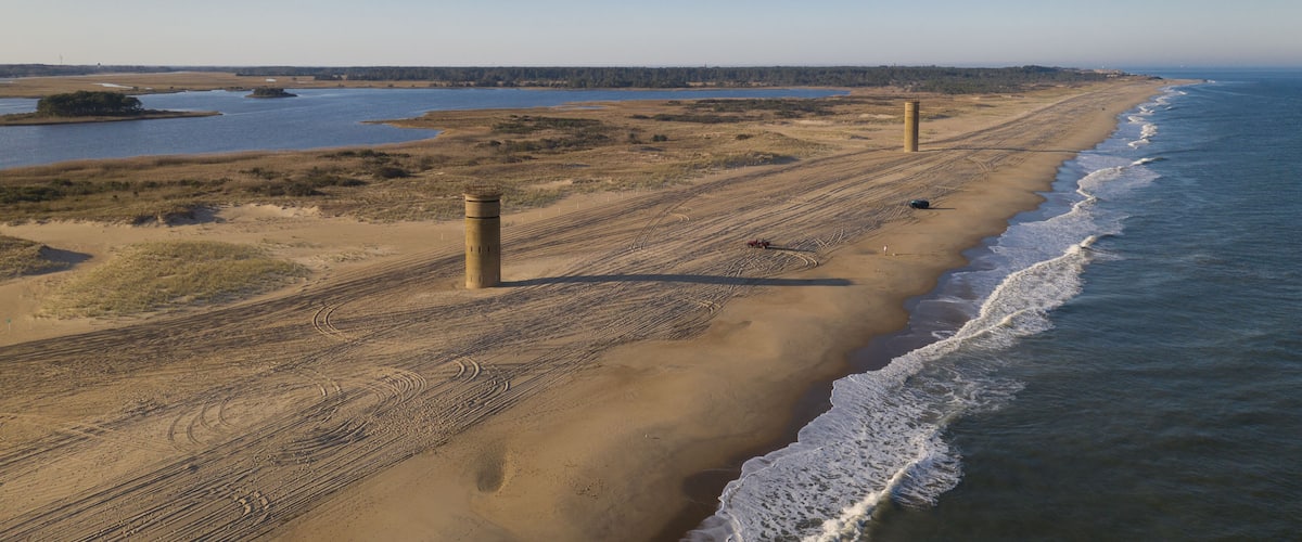 A coastal landscape with a sandy beach, a lighthouse, and a calm sea.
