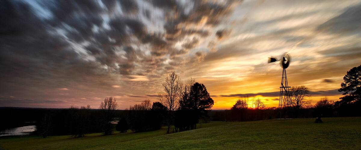 A sunset over a grassy field with a windmill and trees silhouetted against the sky.