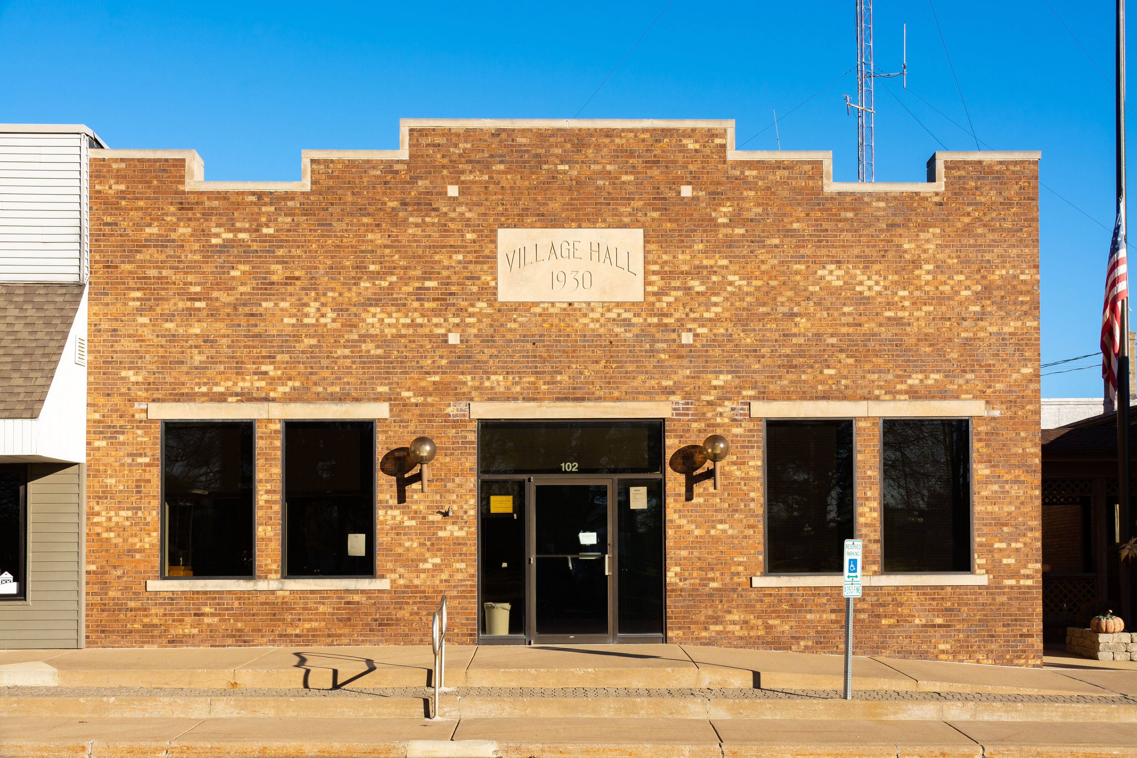 A brick building with a sign reading "Village Hall 1930" and the number "102" above the entrance.