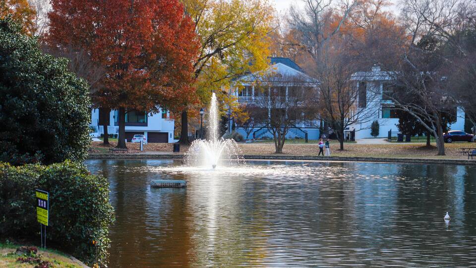 A pond with a fountain, surrounded by autumn-colored trees and a building in the background.