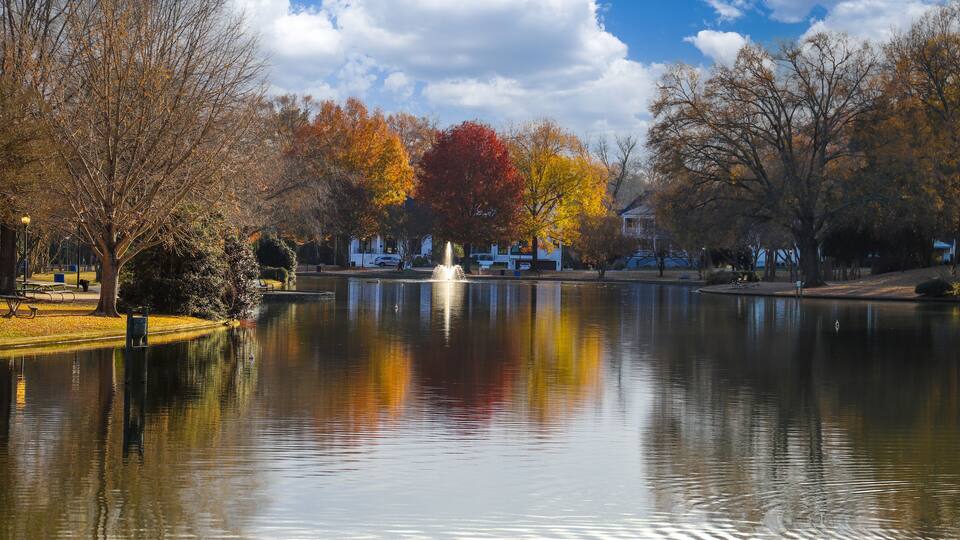 A calm lake with autumn-colored trees, a fountain, and a clear blue sky.