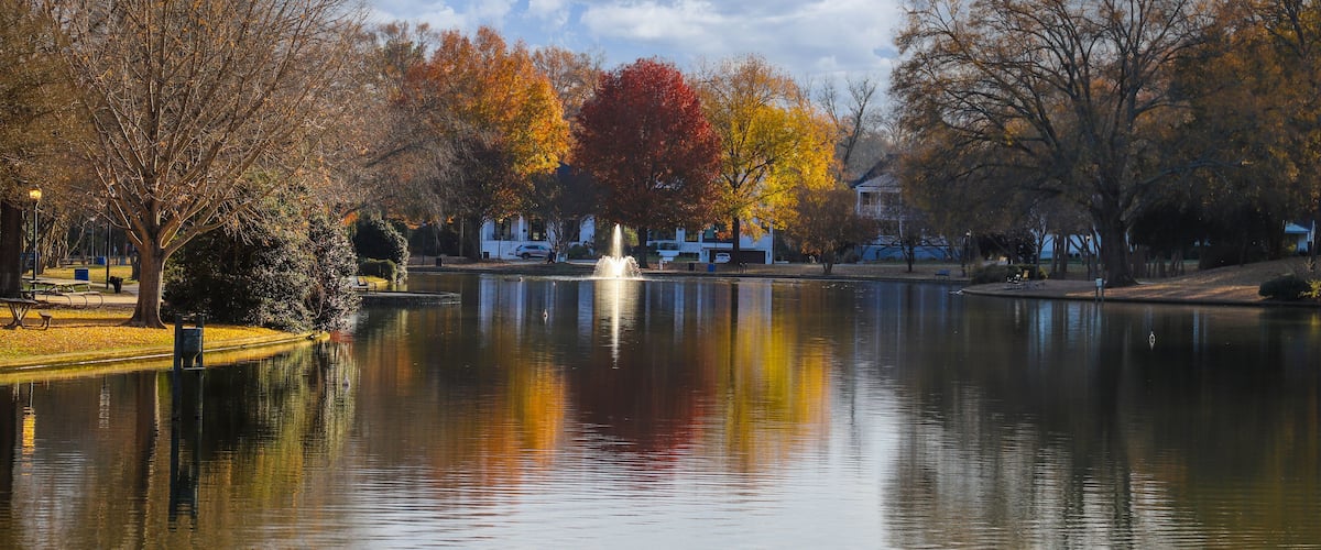A calm lake with autumn-colored trees, a fountain, and a clear blue sky.