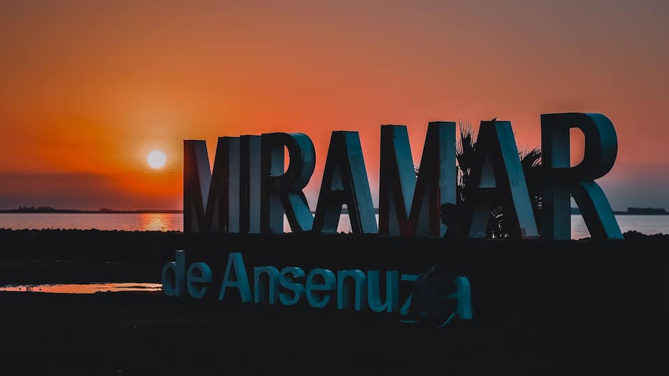 A sunset with the illuminated sign 'MIRAMAR de Anseuz' against a sky of orange and pink hues.