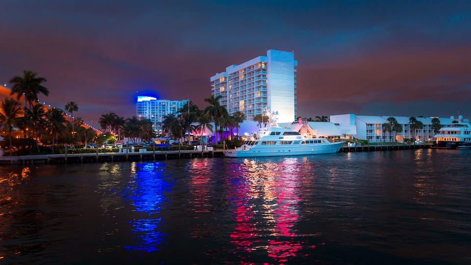 A waterfront cityscape at dusk with a large hotel, palm trees, and a yacht.