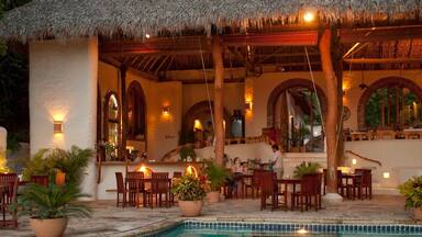 A poolside restaurant with thatched roofs, wooden furniture, and potted plants.