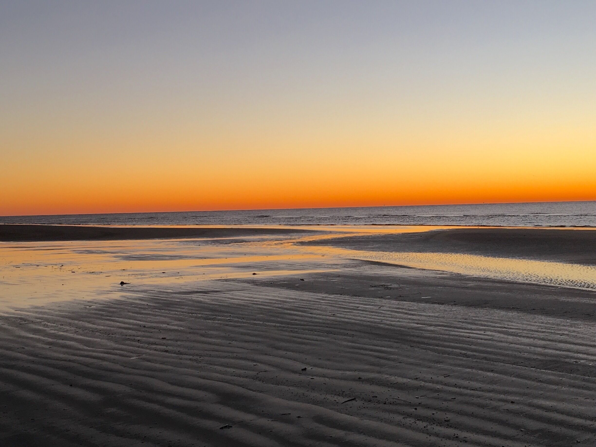 A beach at sunset with sand ripples and a calm sea.