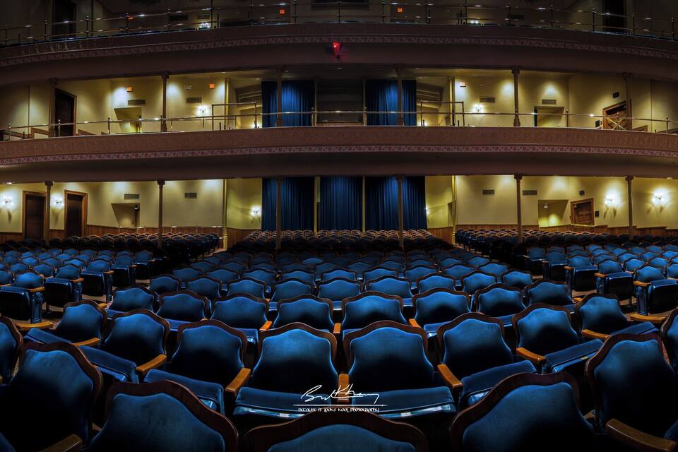 Um teatro com fileiras de assentos azuis, balcões e colunas ricamente decoradas.