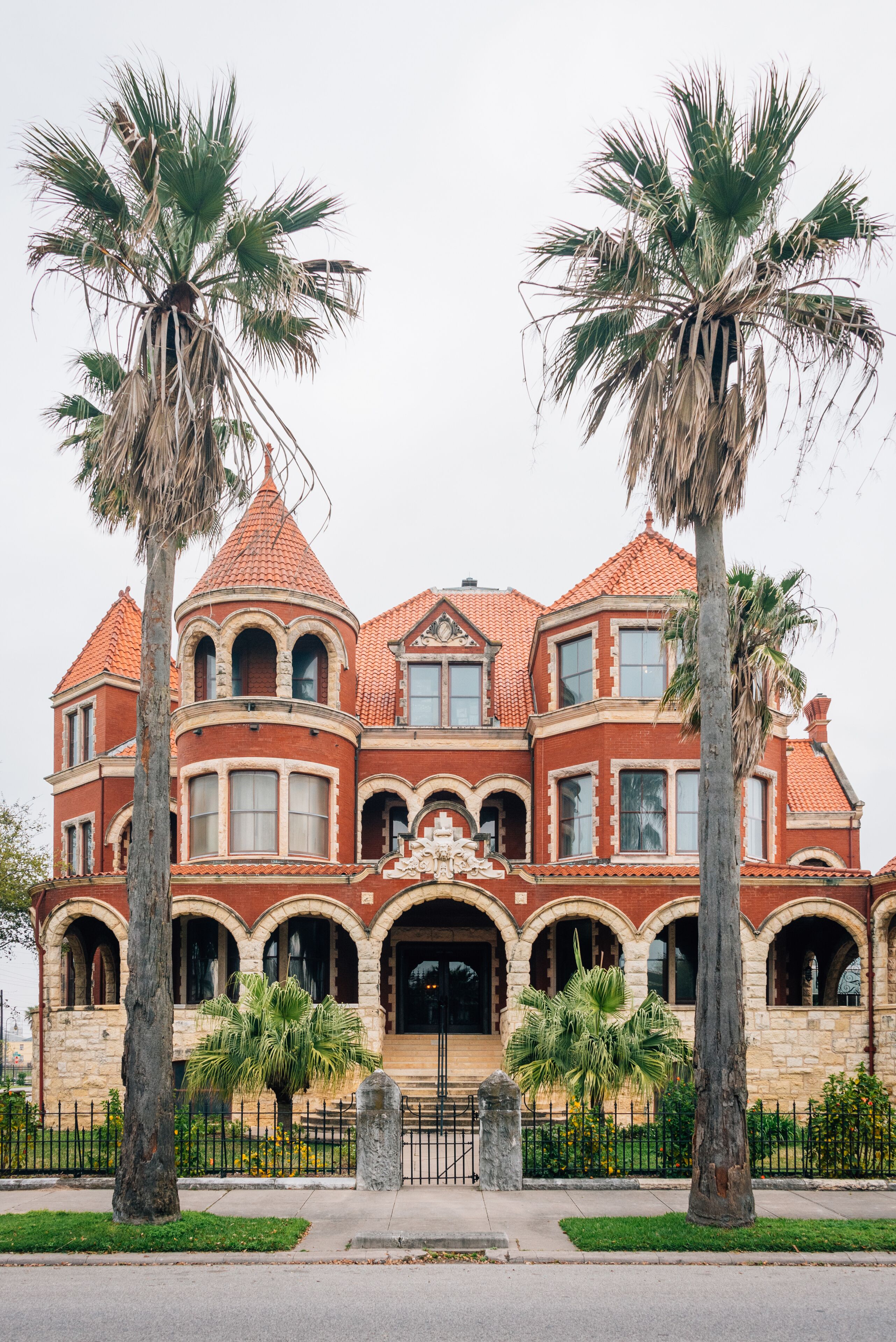 Una gran mansión de ladrillo rojo con ventanas arqueadas y una torre, bordeada de palmeras.