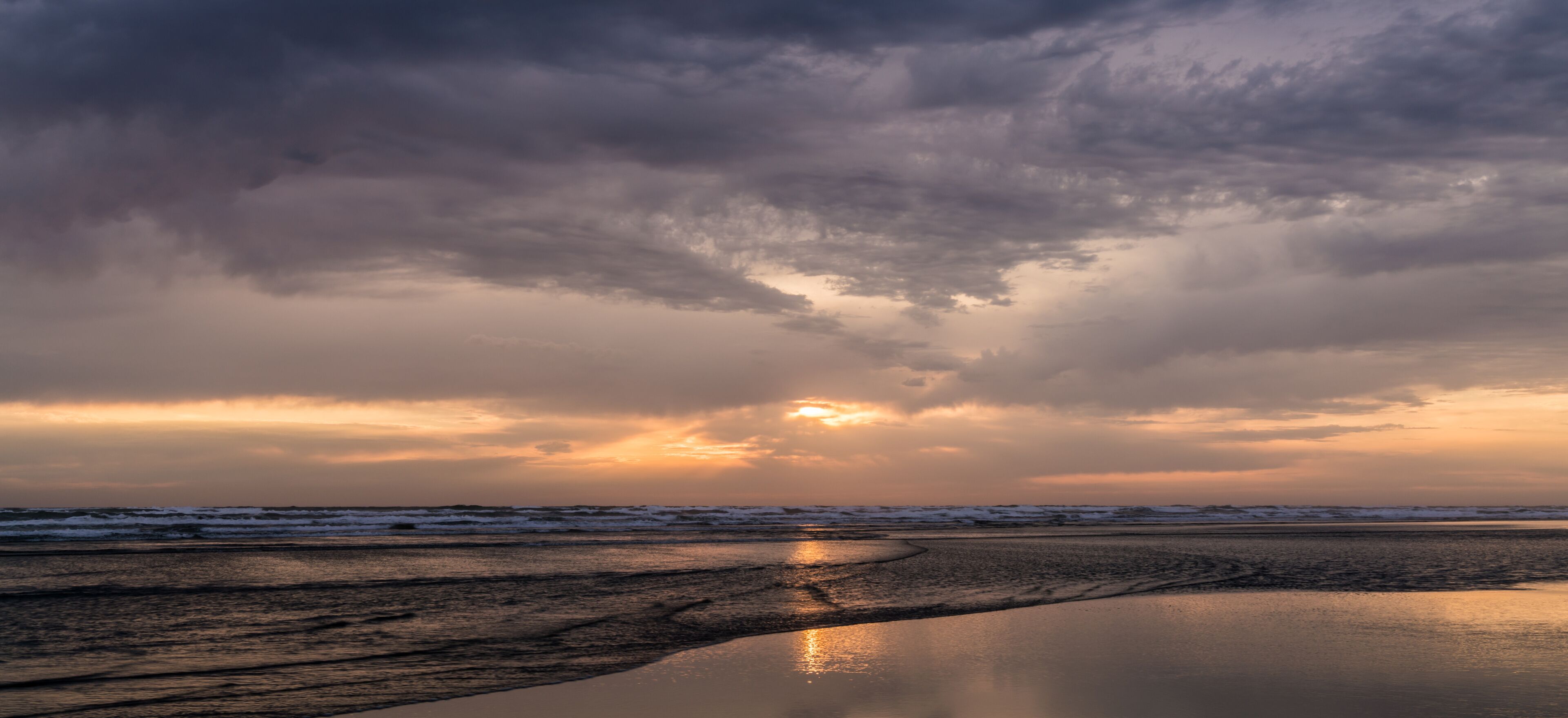 A beach at sunset with a cloudy sky and calm water reflecting the sun.