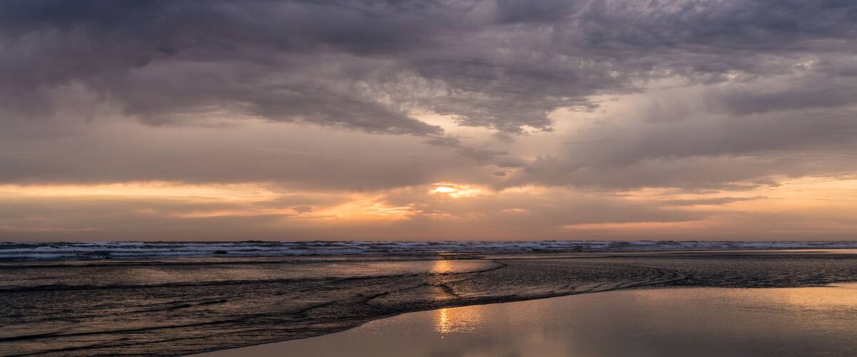 A beach at sunset with a cloudy sky and calm water reflecting the sun.