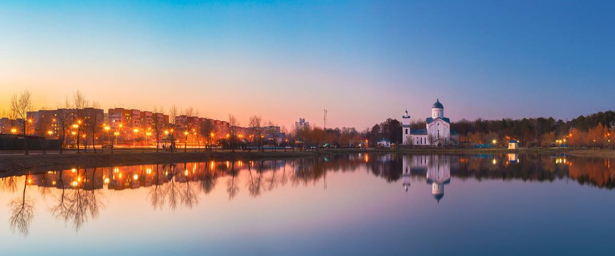 A calm river reflecting buildings and a church with a dome.