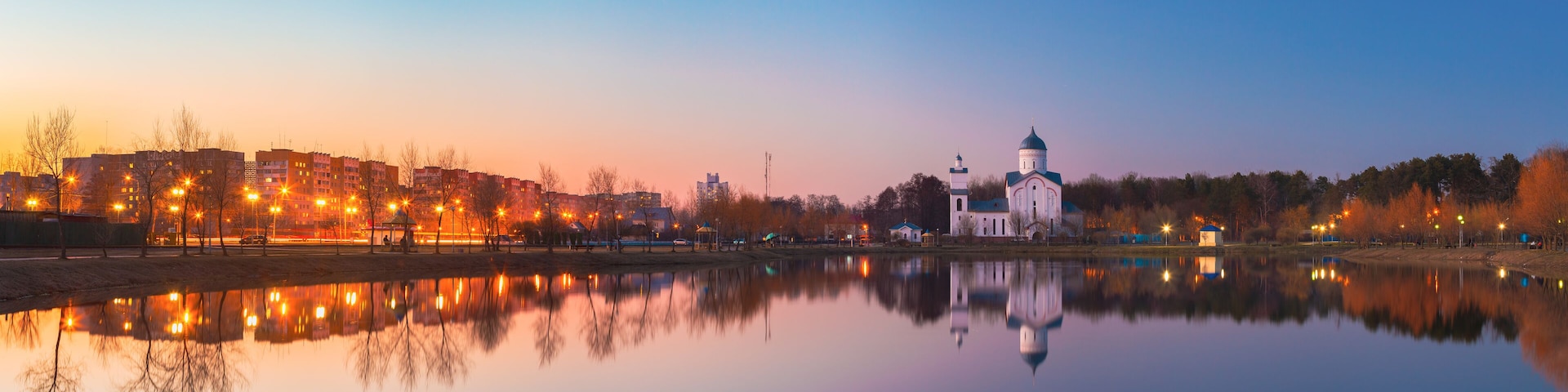 A calm river reflecting buildings and a church with a dome.