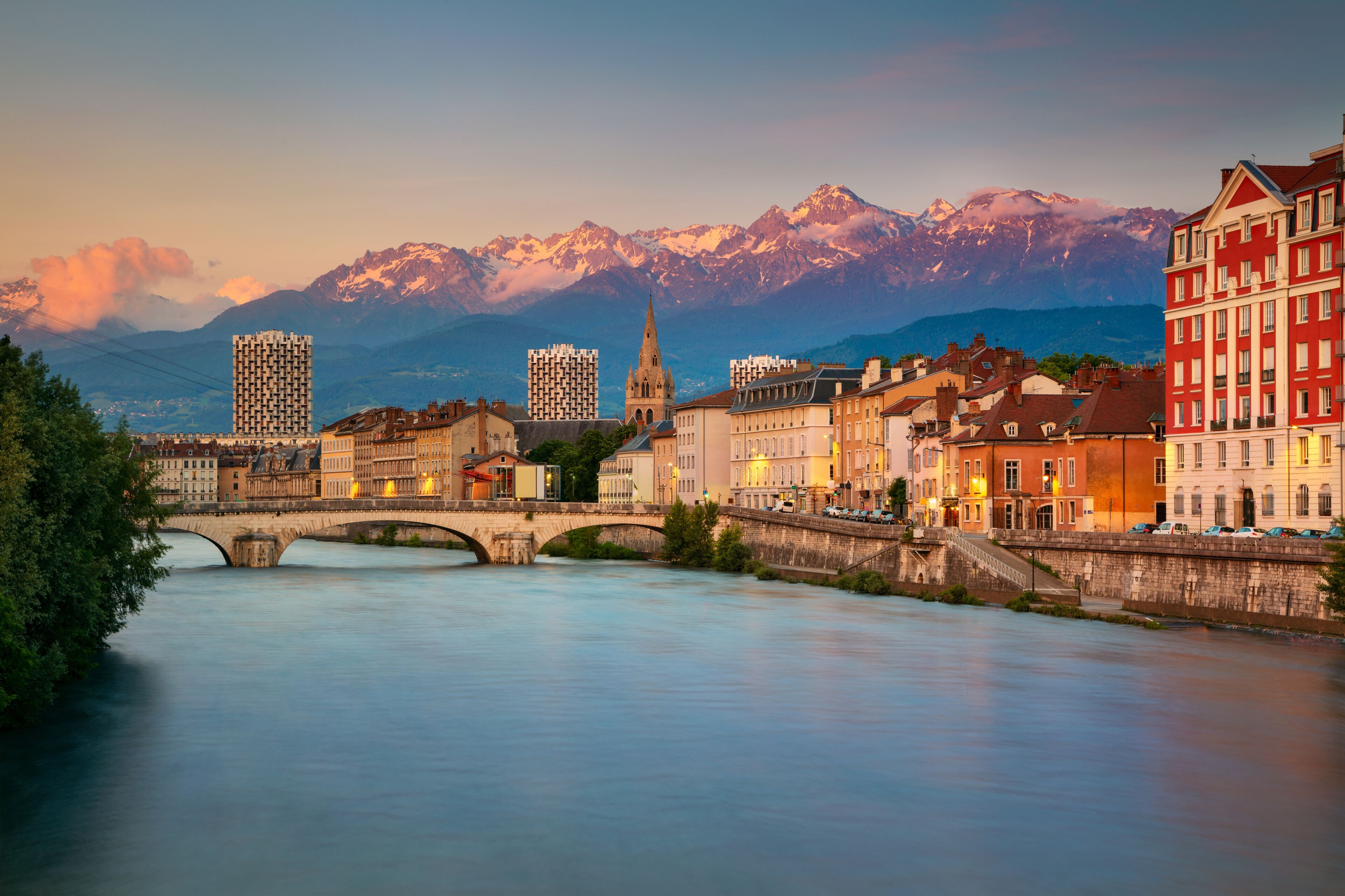 A cityscape with a river, a bridge, and snow-capped mountains in the background.