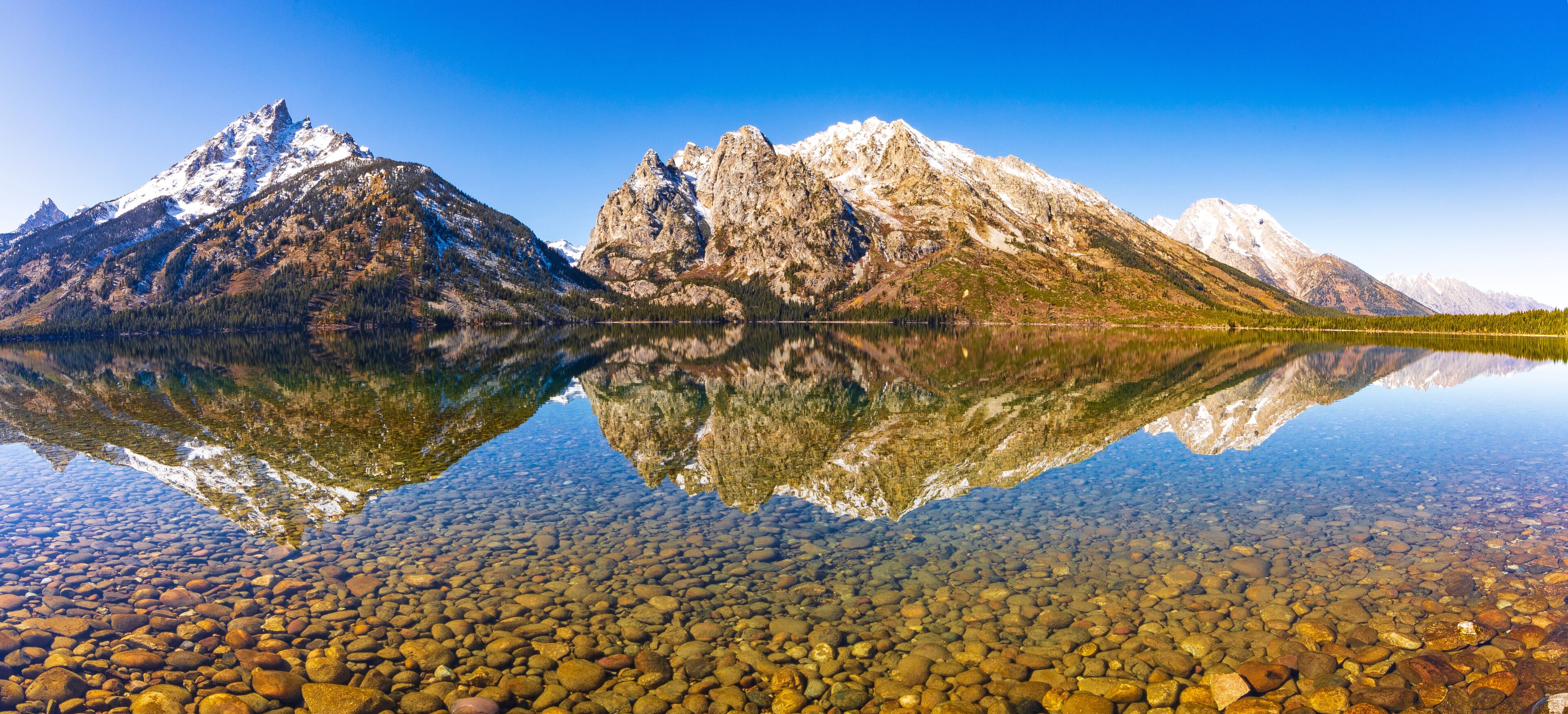 Uma cordilheira refletida em um lago límpido, com o leito pedregoso visível.