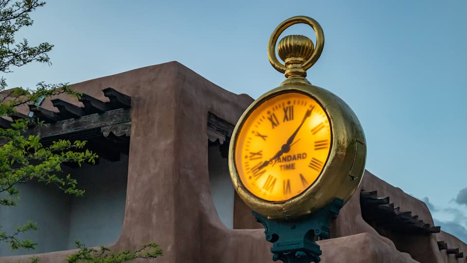A golden clock with Roman numerals set against a building with a brown facade and green foliage.