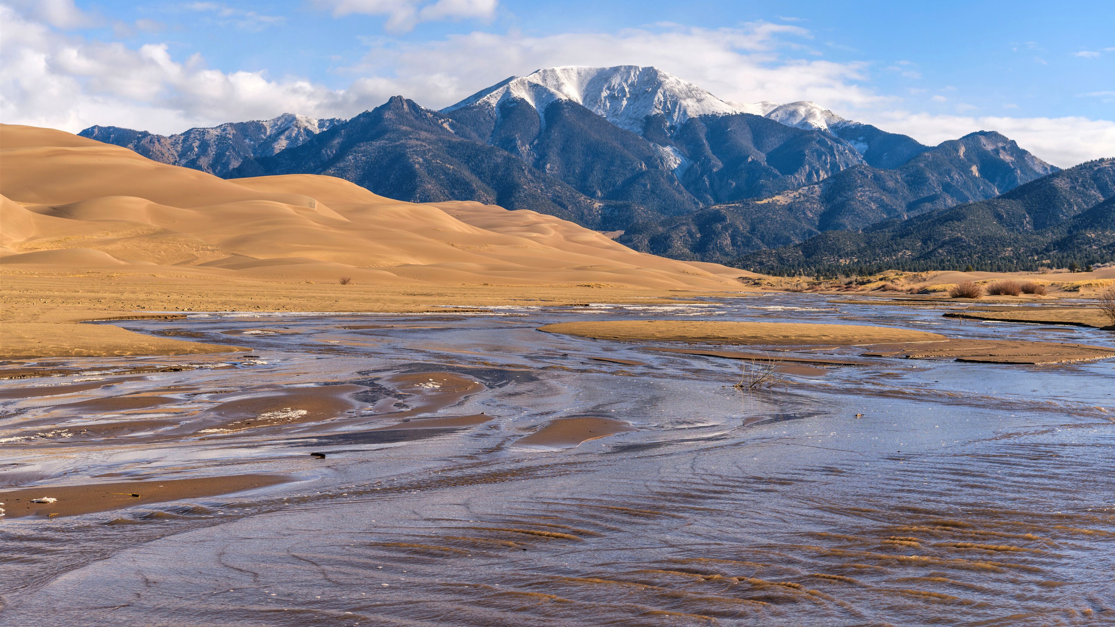 A riverbed with sand dunes and snow-capped mountains in the background.