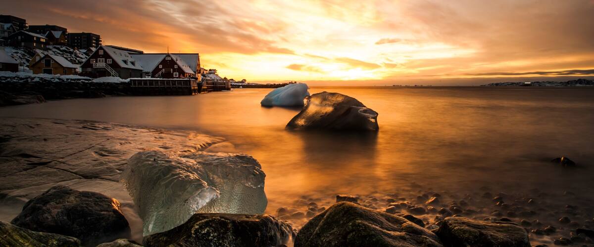 A sunset over a frozen lake with houses on the shore and a large iceberg in the water.