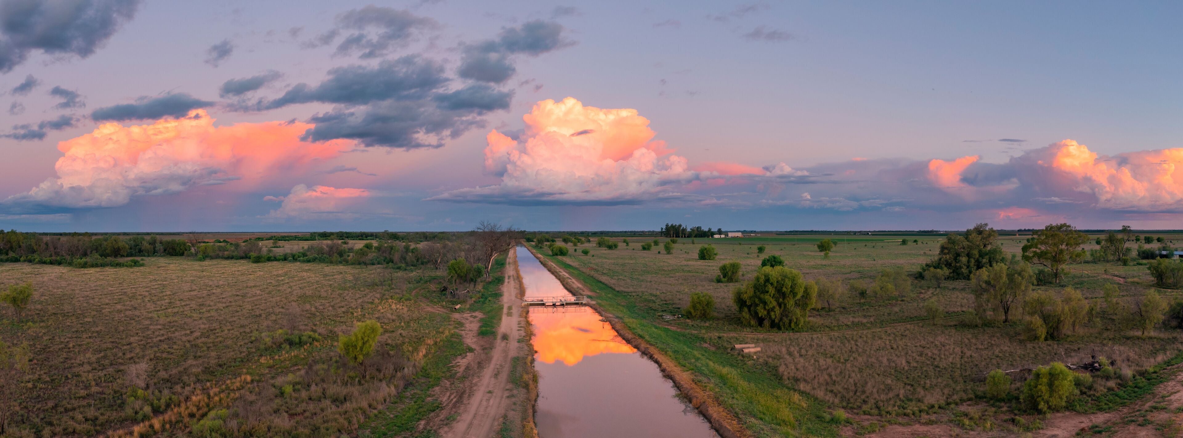 A rural landscape with a dirt road, a water channel, and fields under a sunset sky with large clouds.