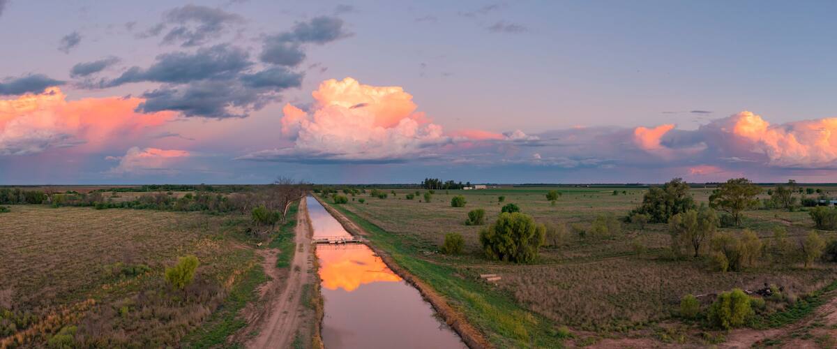 A rural landscape with a dirt road, a water channel, and fields under a sunset sky with large clouds.