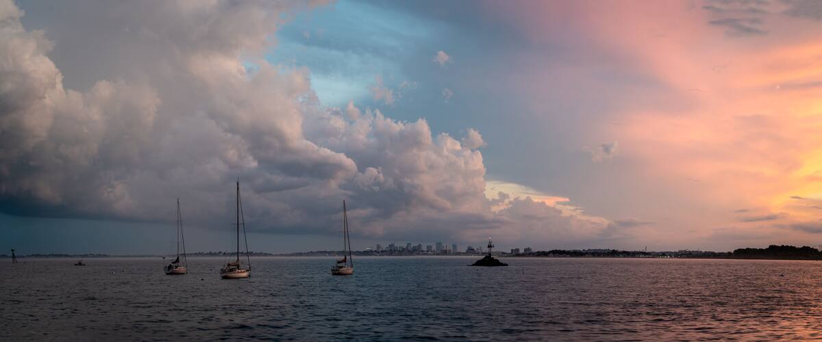 A sunset over a body of water with sailboats and a distant city skyline.