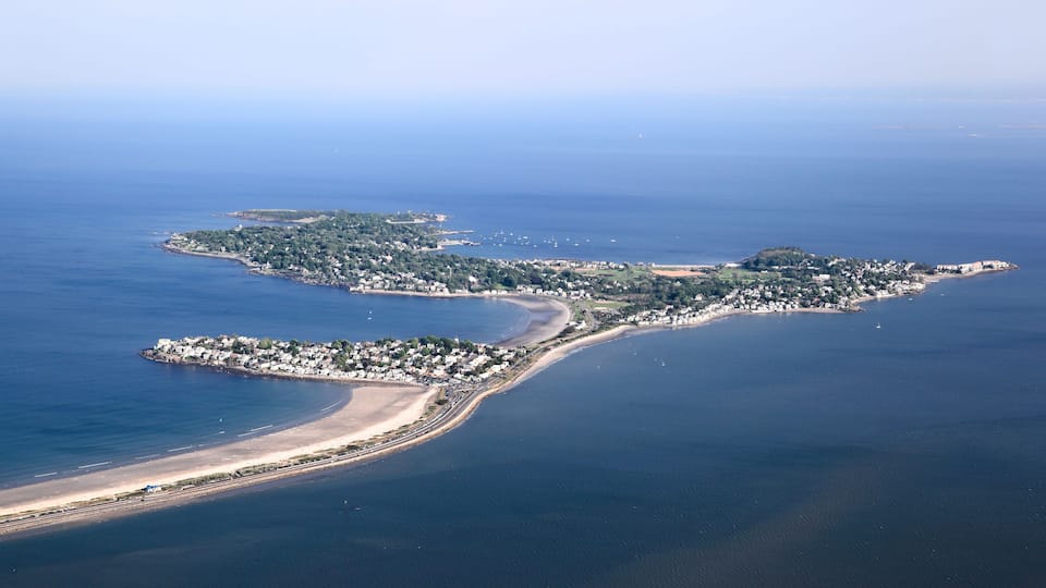 An aerial view of a coastal town with a beach, a marina, and a small island with buildings.