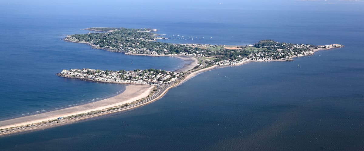 An aerial view of a coastal town with a beach, a marina, and a small island with buildings.
