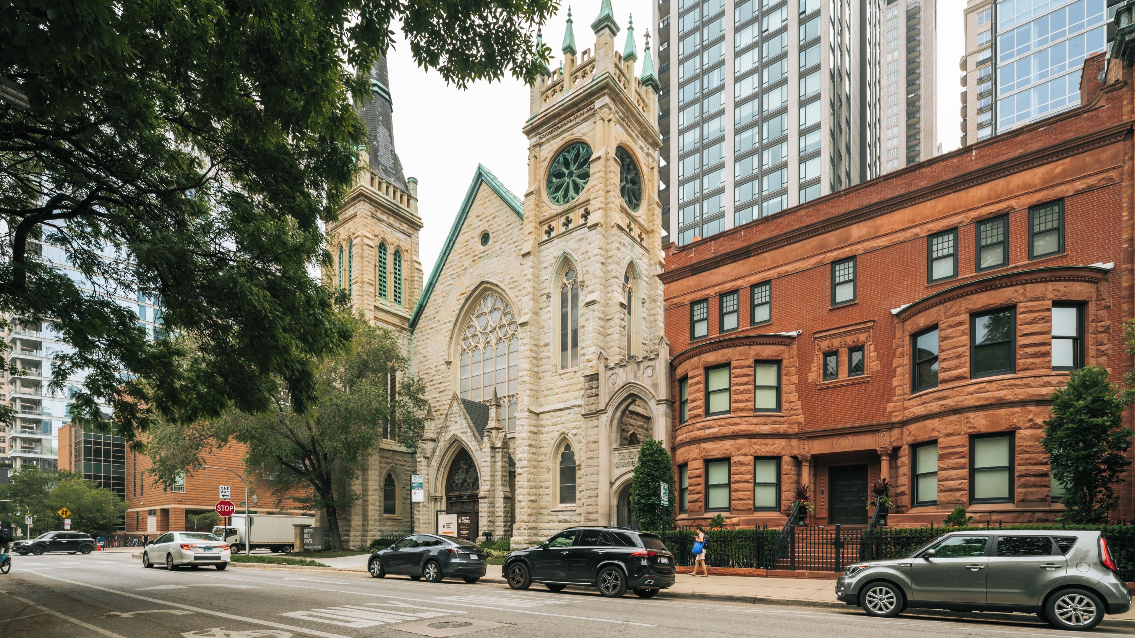 A historic church with a clock tower, surrounded by modern high-rise buildings and parked cars on a city street.