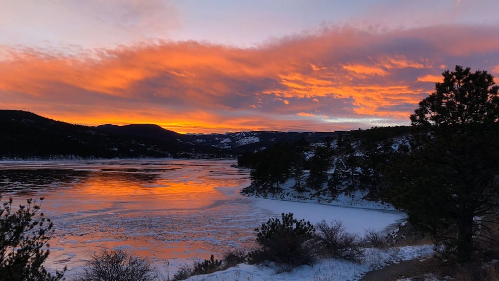Une rivière gelée reflétant les couleurs du coucher de soleil, entourée de collines et d’arbres couverts de neige.