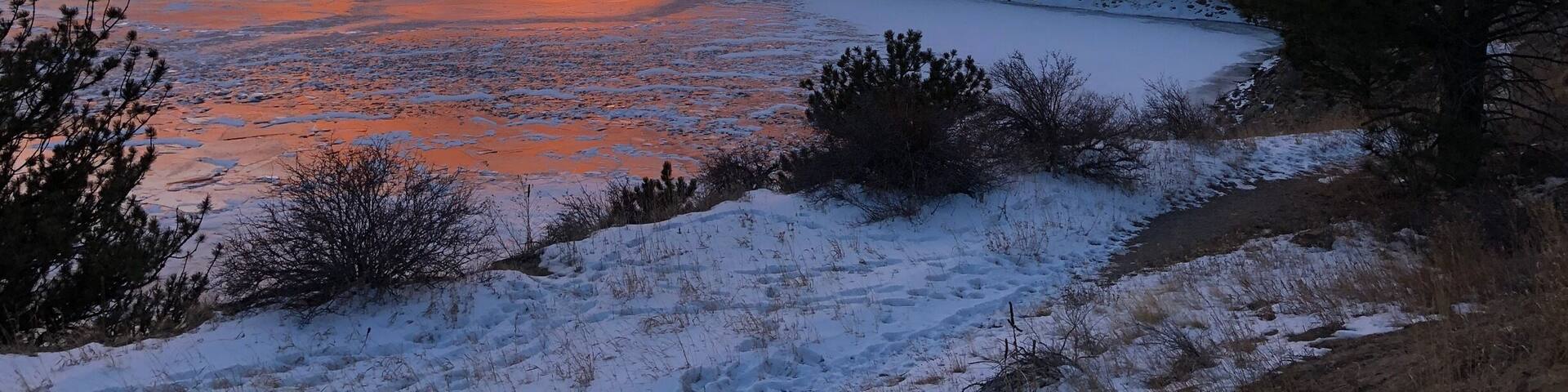 A frozen river reflecting sunset colors, surrounded by snow-covered hills and trees.