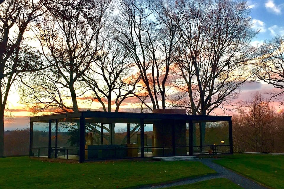 Una estructura moderna de cristal en un parque con árboles y el ocaso al fondo.