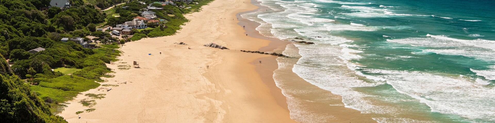 Paisagem costeira com praia arenosa, ondas oceânicas e um morro com vegetação e construções.